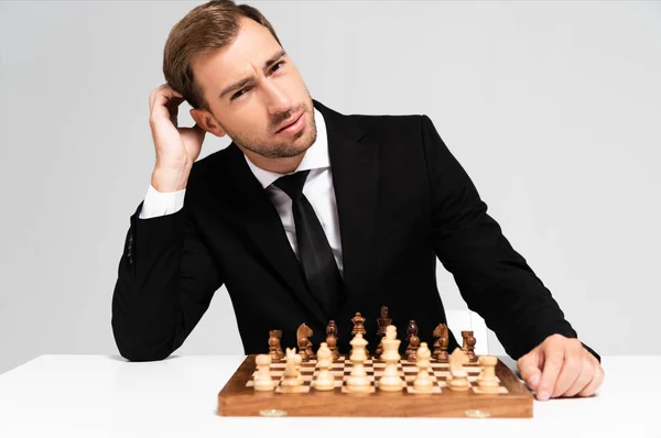Pensativo y guapo hombre de negocios en traje sentado cerca de tablero de ajedrez aislado en gris - foto de stock