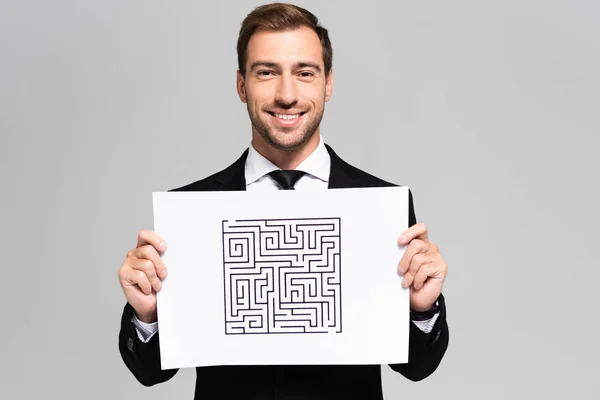 Hombre de negocios sonriente y guapo sosteniendo papel con laberinto aislado en gris - foto de stock