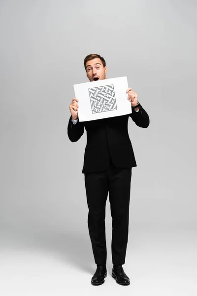 Handsome and shocked businessman in suit holding paper with labyrinth on grey background — Stock Photo