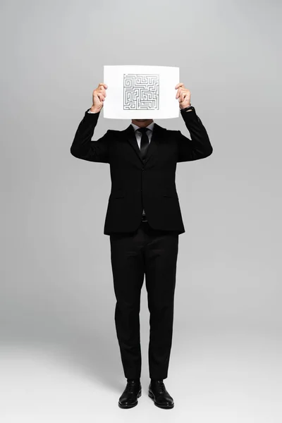 Empresário obscurecendo rosto com papel com labirinto sobre fundo cinza — Fotografia de Stock