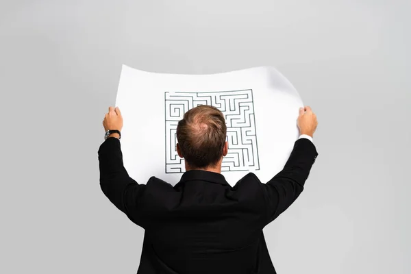 Vue arrière de l'homme d'affaires en costume regardant le papier avec labyrinthe isolé sur gris — Photo de stock