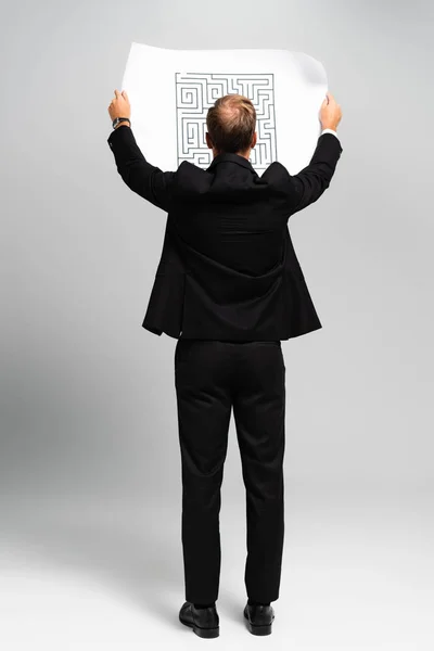 Back view of businessman in suit looking at paper with labyrinth on grey background — Stock Photo