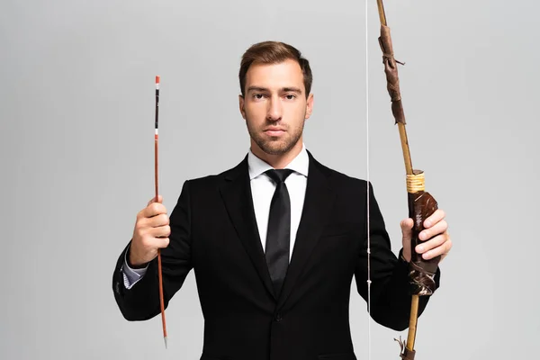 Guapo hombre de negocios en traje sosteniendo arco y flecha aislado en gris - foto de stock