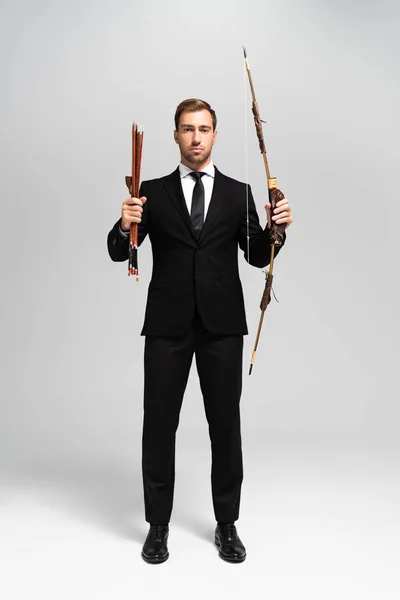 Guapo hombre de negocios en traje sosteniendo arco y flechas sobre fondo gris - foto de stock