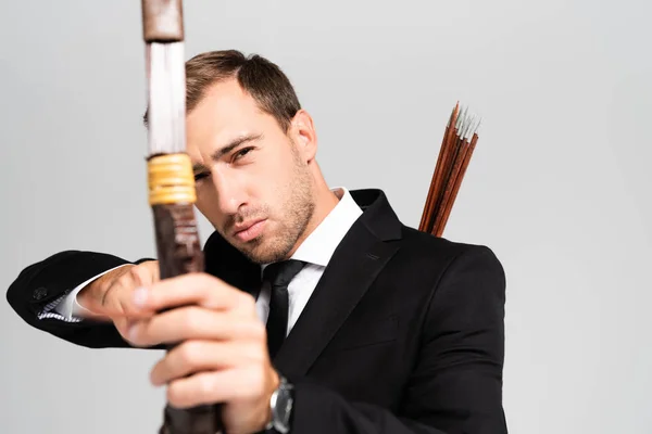 Selective focus of handsome businessman in suit holding bow and shooting with arrow isolated on grey — Stock Photo
