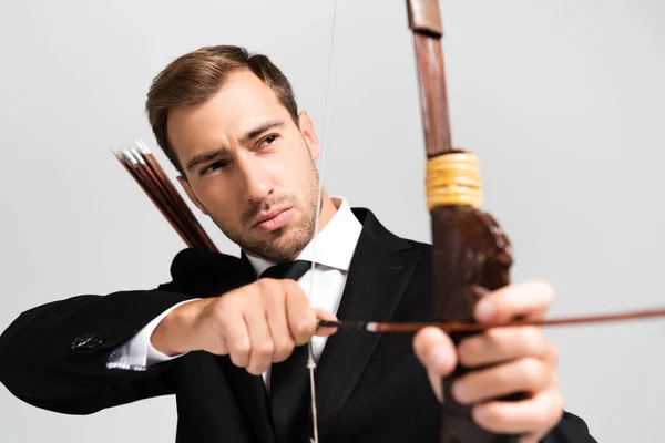 Selektiver Fokus eines gutaussehenden Geschäftsmannes im Anzug, der Bogen hält und mit Pfeil auf grau isoliert schießt — Stockfoto