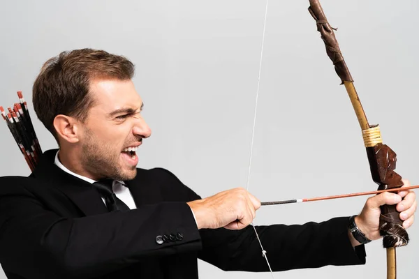 Side view of angry businessman in suit holding bow and shooting with arrow isolated on grey — Stock Photo