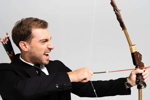 Seitenansicht eines wütenden Geschäftsmannes im Anzug, der Bogen hält und mit Pfeil auf grau isoliert schießt — Stockfoto