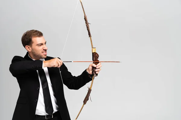 Hombre de negocios enojado en traje sosteniendo el arco y disparando con flecha aislada en gris - foto de stock