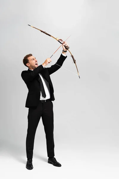 Сердитий бізнесмен в костюмі тримає лук і стріляє зі стрілою на сірому фоні — стокове фото