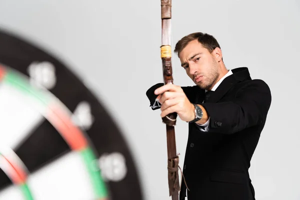 Selective focus of handsome businessman in suit holding bow and shooting at target isolated on grey — Stock Photo