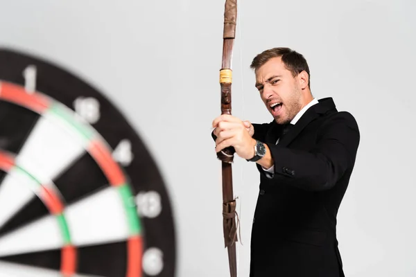 Selective focus of angry businessman in suit holding bow and shooting at target isolated on grey — Stock Photo