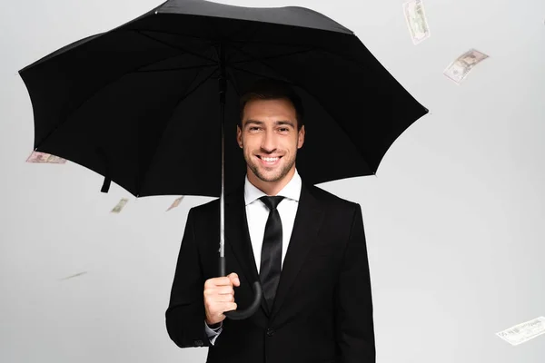 Hombre de negocios guapo y sonriente en traje con paraguas y de pie cerca de la caída de billetes de dólar aislados en gris - foto de stock