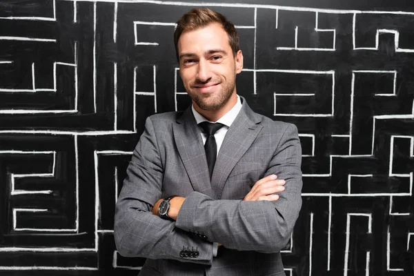 Handsome and smiling businessman in suit with crossed arms standing near labyrinth — Stock Photo