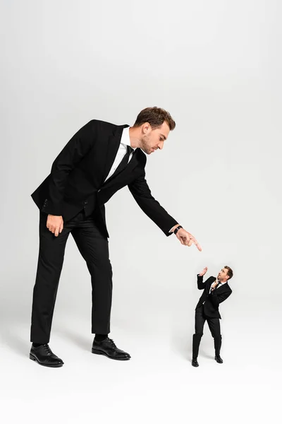 Hombre de negocios enojado en traje señalando con el dedo a la marioneta asustada sobre fondo gris - foto de stock