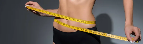 Cropped view of slim woman in underwear holding measuring tape on waist on dark background, panoramic shot — Stock Photo