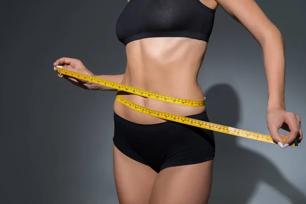 Cropped view of slim woman in underwear holding measuring tape on waist on dark background — Stock Photo