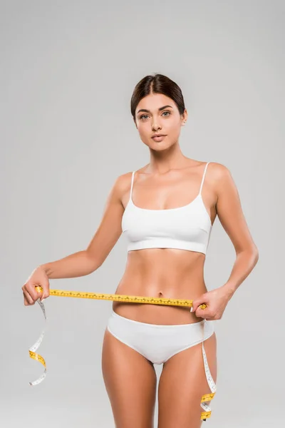 Beautiful slim woman in underwear holding measuring tape on waist isolated on grey — Stock Photo