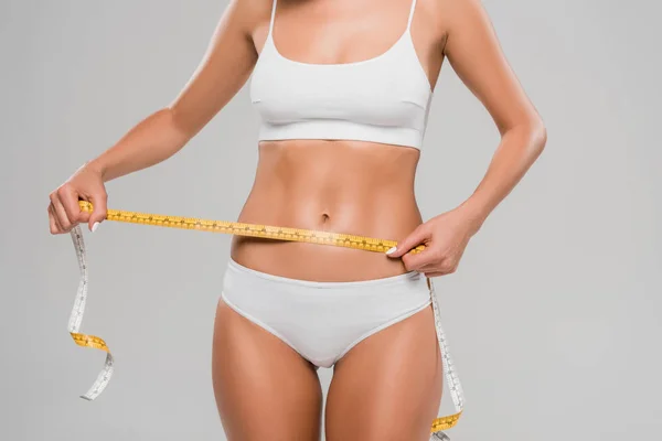 Partial view of beautiful slim woman in underwear holding measuring tape on waist isolated on grey — Stock Photo