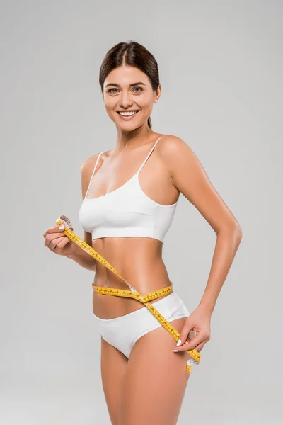 Happy beautiful slim woman in underwear holding measuring tape on waist isolated on grey — Stock Photo