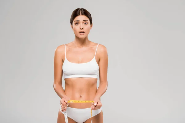 Shocked beautiful slim woman in underwear holding measuring tape isolated on grey — Stock Photo