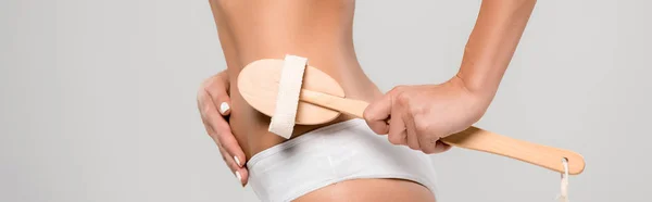 Vue recadrée d'une femme mince en sous-vêtements massant le corps avec une brosse de massage en bois isolée sur fond gris, panoramique — Photo de stock