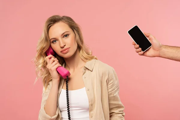 Mujer rubia hablando por teléfono y la mano sosteniendo teléfono inteligente aislado en rosa - foto de stock