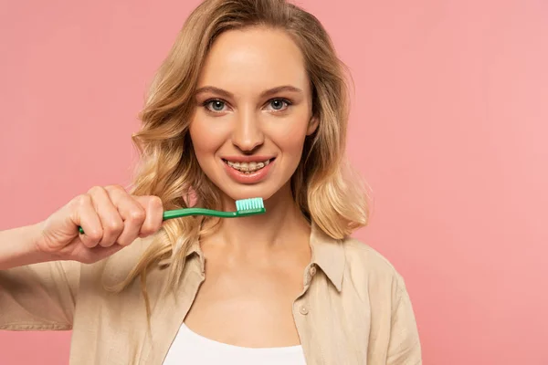 Femme blonde souriante avec des appareils dentaires tenant la brosse à dents isolée sur rose — Photo de stock