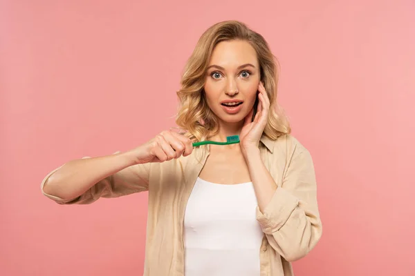 Femme blonde choquée tenant une brosse à dents isolée sur rose — Photo de stock