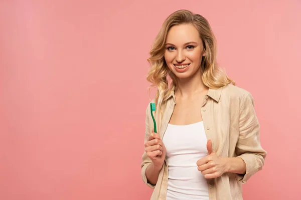 Jeune femme souriante tenant une brosse à dents et montrant le pouce isolé sur rose — Photo de stock