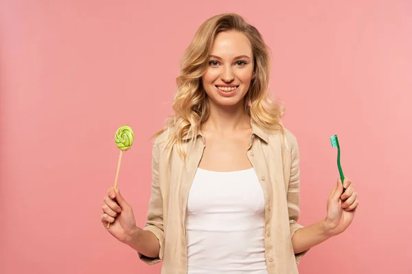 Smiling blonde woman holding toothbrush and lollipop isolated on pink — Stock Photo