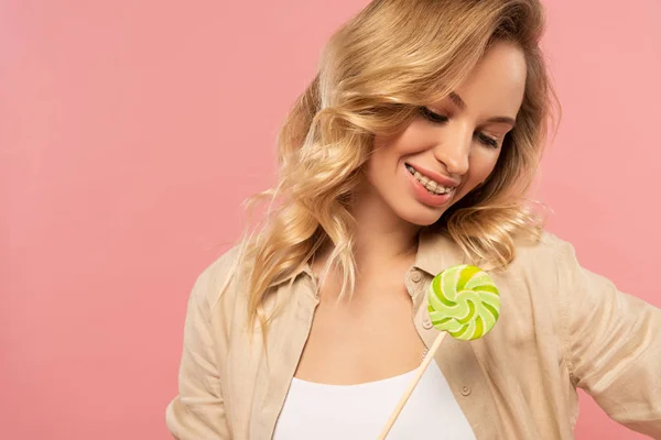 Mujer rubia sonriente mirando piruleta aislada en rosa - foto de stock