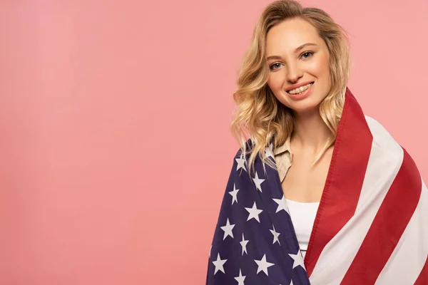 Jeune femme souriante enveloppée dans un drapeau américain isolé sur rose — Photo de stock