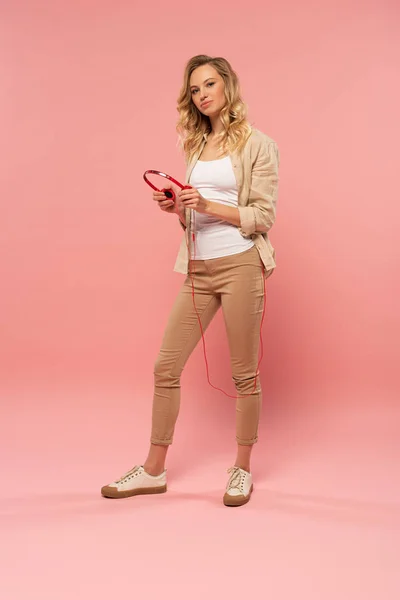 Mulher atraente segurando fones de ouvido no fundo rosa — Fotografia de Stock