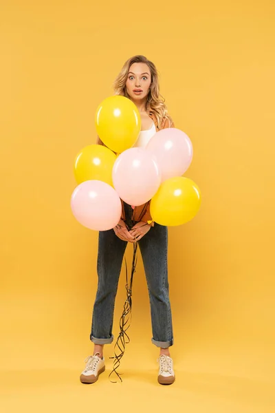 Mujer rubia sorprendida sosteniendo globos sobre fondo amarillo - foto de stock