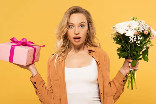 Schockierte blonde Frau hält Blumenstrauß und Geschenkbox isoliert auf gelb — Stockfoto