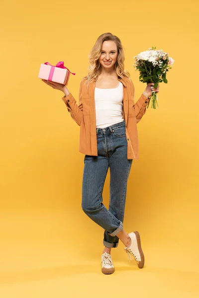 Lächelnde blonde Frau mit Blumenstrauß und Geschenkbox auf gelbem Hintergrund — Stockfoto