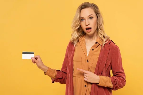 Schockierte blonde Frau zeigt auf Kreditkarte isoliert auf gelb — Stockfoto