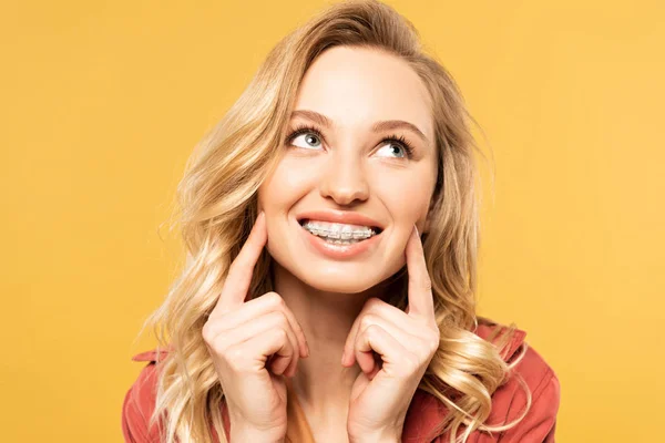 Mujer rubia sonriente con frenos dentales mirando hacia arriba aislado en amarillo - foto de stock