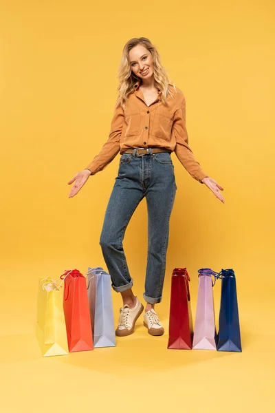 Mujer rubia sonriente de pie junto a las bolsas de la compra en el suelo sobre fondo amarillo - foto de stock