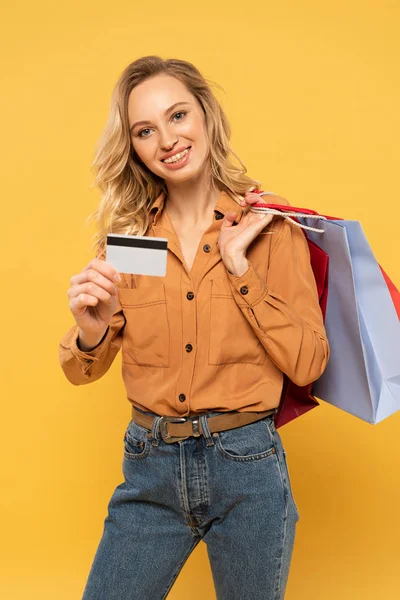 Donna sorridente mostrando carta di credito e tenendo borse della spesa isolate sul giallo — Foto stock