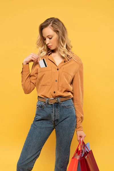 Blonde woman putting credit card in shirt pocket and holding shopping bags isolated on yellow — Stock Photo