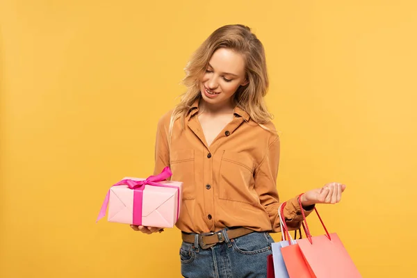 Lächelnde Frau blickt auf Geschenkbox und hält Einkaufstüten isoliert auf gelb — Stockfoto