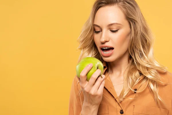 Mujer joven con frenos dentales mordiendo manzana verde aislada en amarillo - foto de stock