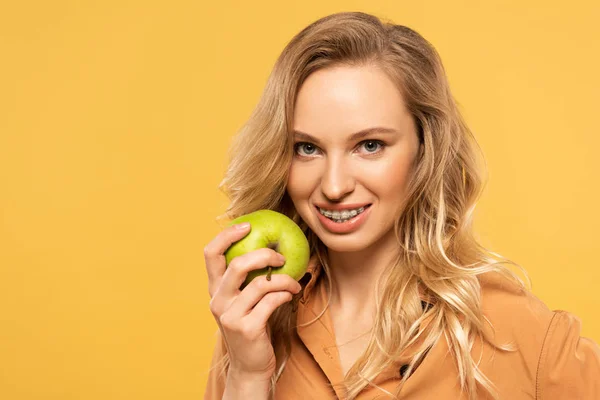 Lächelnde blonde Frau mit Zahnspange, die grünen Apfel isoliert auf gelb hält — Stockfoto