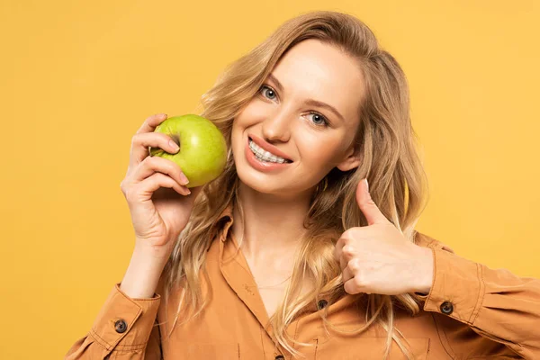 Усміхнена жінка тримає зелене яблуко і показує жест великого пальця ізольовано на жовтому — стокове фото