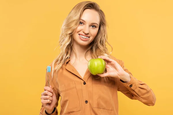 Усміхнена жінка тримає зелене яблуко і зубну щітку ізольовано на жовтому — стокове фото