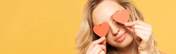 Smiling woman covering eyes with paper heart signs isolated on yellow, panoramic shot — Stock Photo