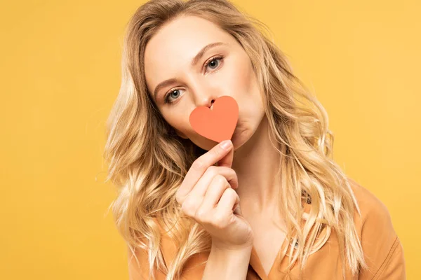 Femme blonde couvrant la bouche avec du papier signe cardiaque isolé sur jaune — Photo de stock