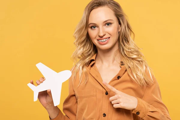 Lächelnde blonde Frau zeigt auf Papierflugzeug isoliert auf gelb — Stockfoto